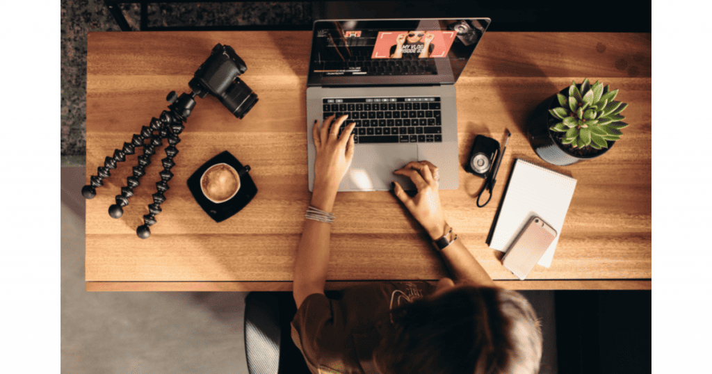 Vue de dessus d'une personne tapant sur un ordinateur portable, avec un mini trépied et une caméra, une tasse de café, un smartphone et une plante sur un bureau en bois, illustrant la production de vidéos courtes pour les réseaux sociaux - Digitandco.
