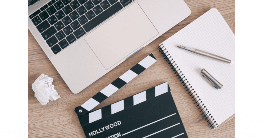 Un ordinateur portable, un clap de cinéma "Hollywood", un carnet à spirale avec un stylo, et une boule de papier froissé sur un bureau en bois, symbolisant la création de contenu vidéo court pour les réseaux sociaux - Digitandco.
