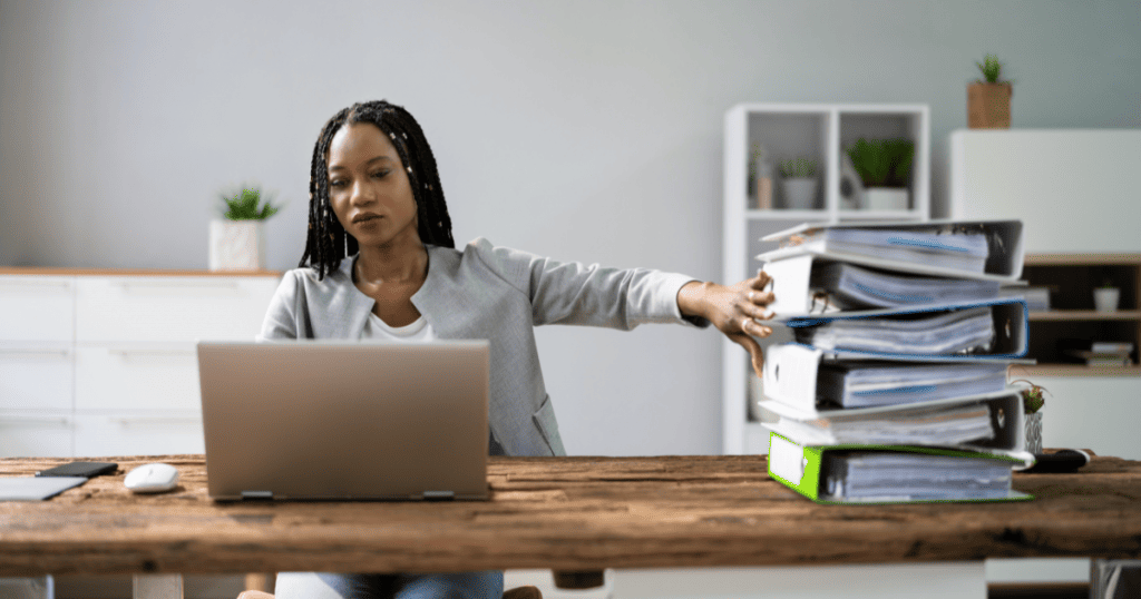 Une femme assise à un bureau, travaillant sur son ordinateur portable tout en éloignant d'un geste de la main une pile de classeurs, symbolisant le passage de la gestion administrative papier à la numérisation - Digitandco.