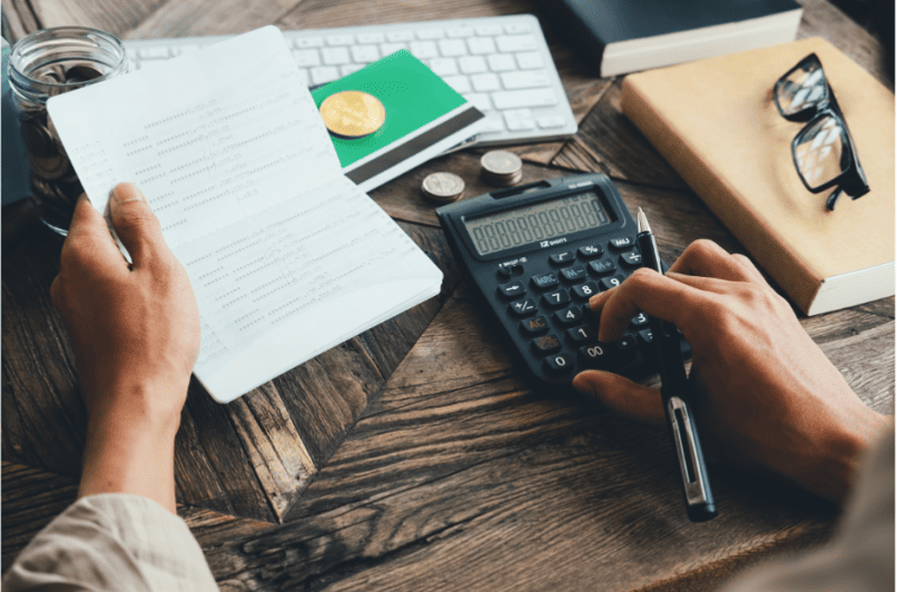 Une personne utilisant une calculatrice et tenant un livret de comptes ou un registre financier, avec des pièces de monnaie, des lunettes et des livres sur un bureau en bois, illustrant la gestion comptable et financière - Digitandco