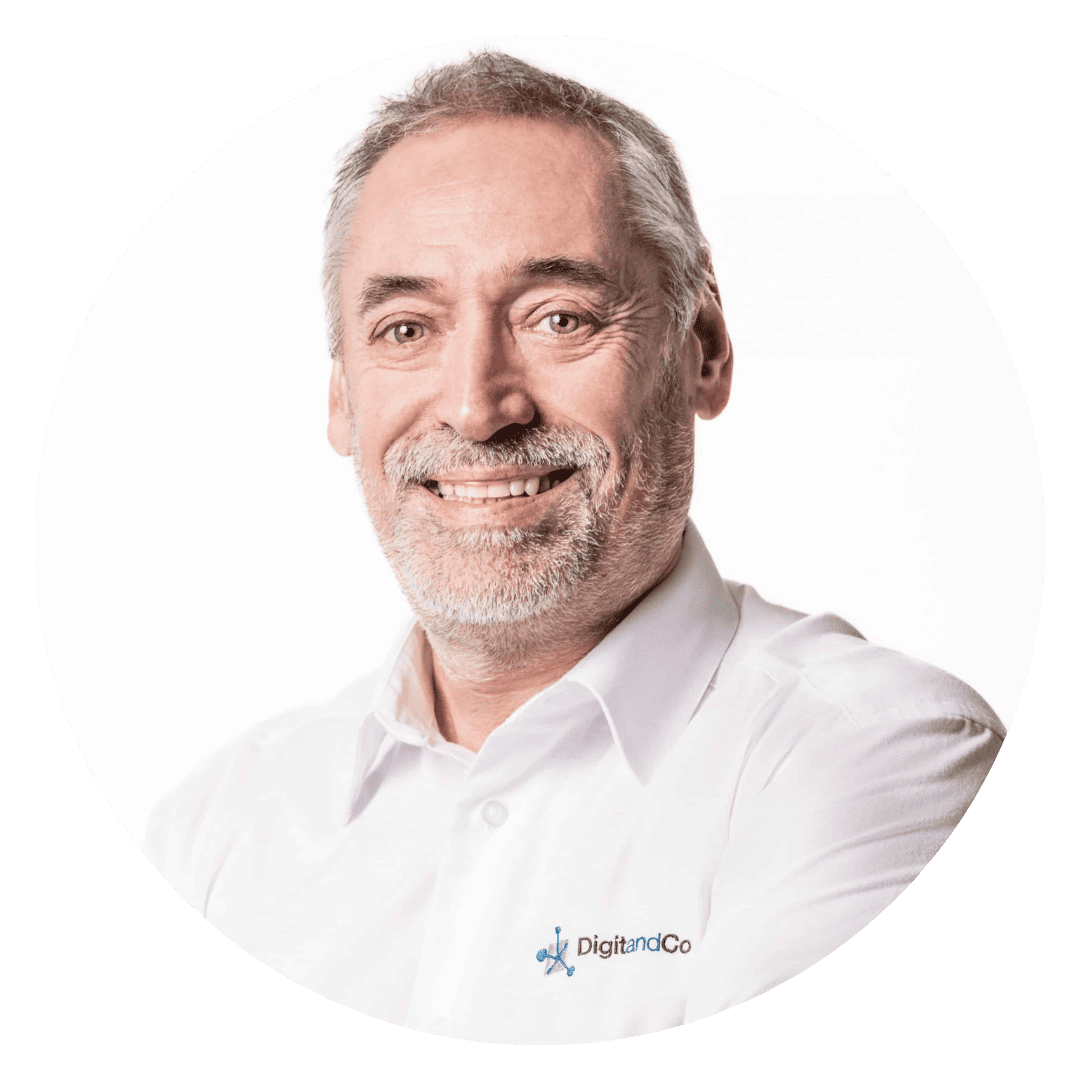 Portrait de Benoît Bornet, fondateur de Digitandco, un homme souriant aux cheveux grisonnants et à la barbe, vêtu d'une chemise blanche sur laquelle est brodé le logo de l'entreprise - Digitandco.