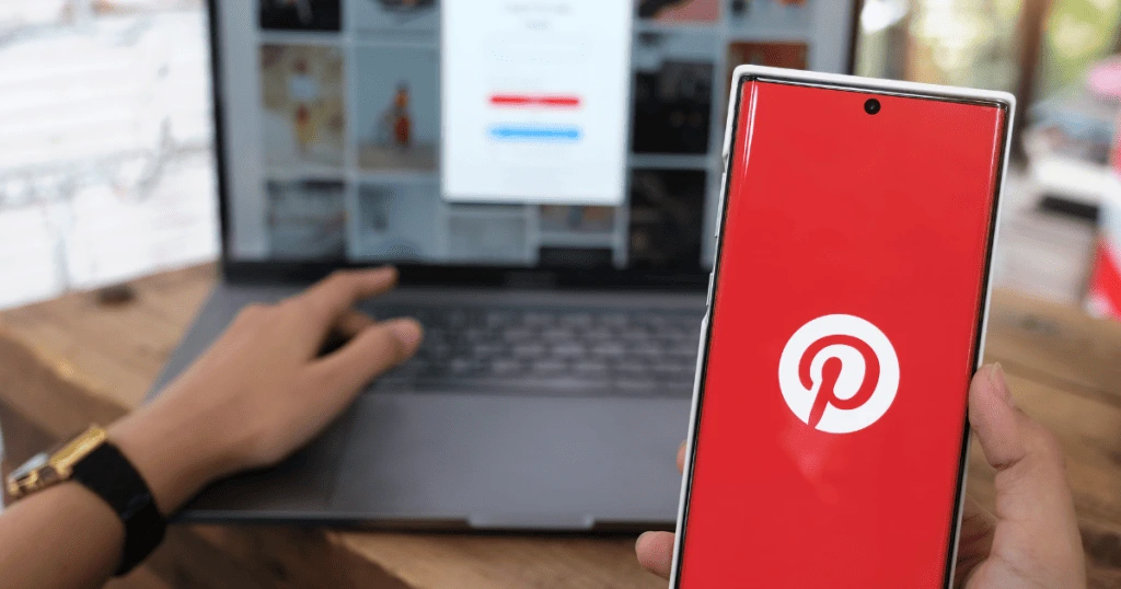 Une personne tenant un smartphone avec l'application Pinterest affichée à l'écran, un ordinateur portable ouvert en arrière-plan montrant une page web de la plateforme, illustrant l'utilisation de Pinterest sur mobile et ordinateur.