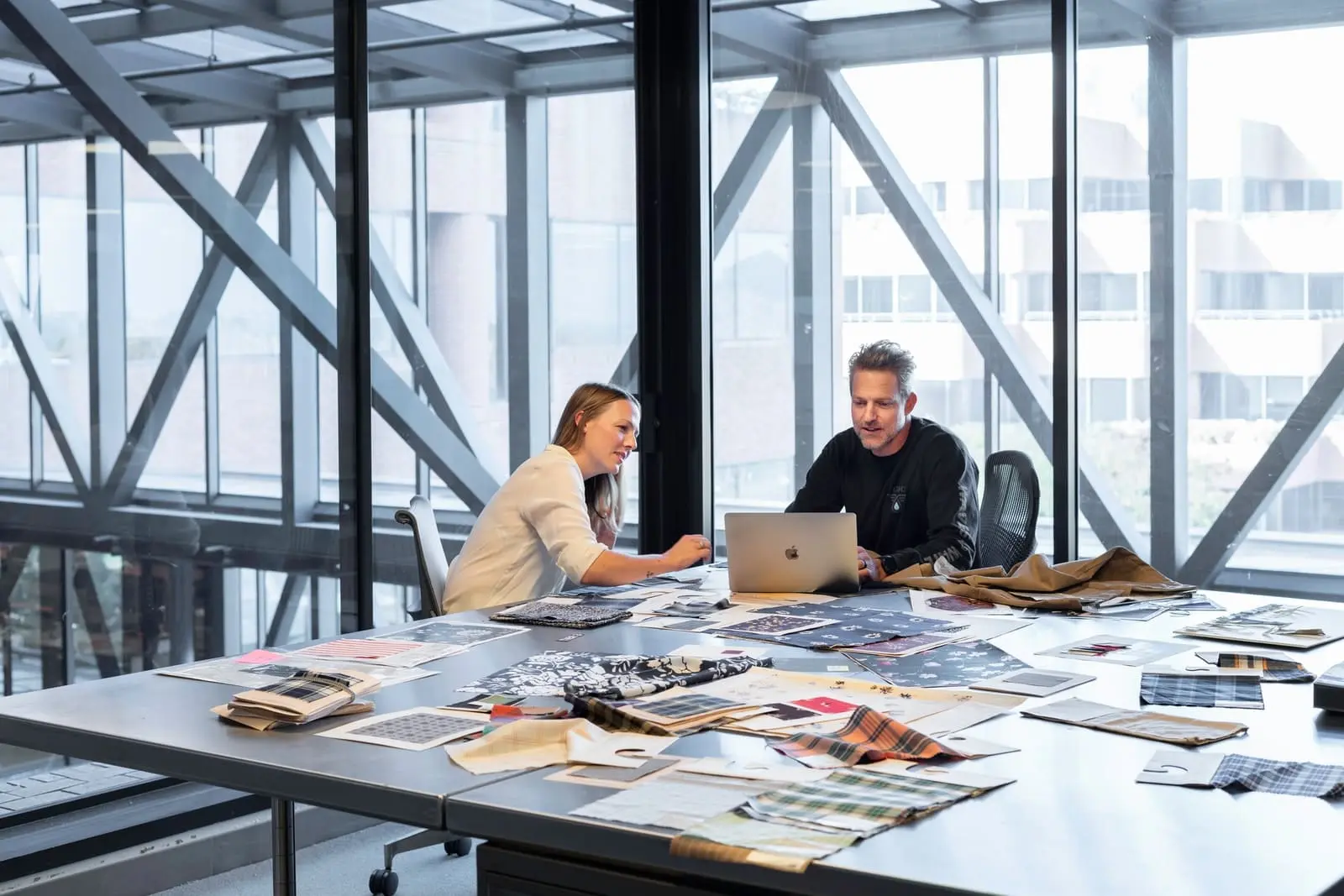 Deux professionnels collaborant autour d'un ordinateur portable, entourés de documents et d'échantillons, symbolisant la mise en valeur des expériences, des réalisations et des compétences sur un profil LinkedIn.