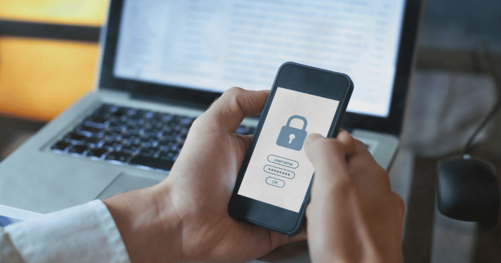 Une personne tenant un smartphone qui affiche un écran de connexion avec un cadenas, symbolisant la sécurité des données sensibles sur les appareils mobiles et la protection contre l'accès non autorisé. Un ordinateur portable est en arrière-plan - Digitandco.
